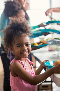 Cute African American preschool age girl smiles while receiving a healthy meal at a local soup kitchen. Her family is in the line behind her also receiving a meal.